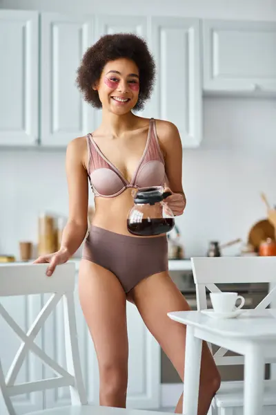 Curly happy african american woman with eye patches standing in underwear with coffee pot — Stock Photo