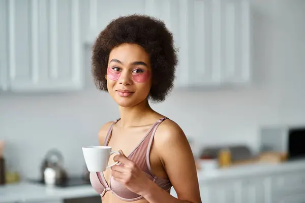 Mujer afroamericana bonita con parches en los ojos de pie en ropa interior y disfrutar del café en la taza - foto de stock