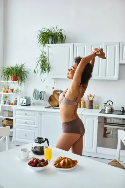 Sorridente donna africana americana in lingerie che si estende vicino a deliziosa colazione sul tavolo della cucina — Foto stock