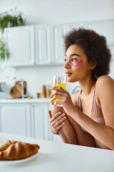 Femme africaine américaine rêveuse en lingerie boire du jus d'orange près du petit déjeuner sur la table de cuisine — Photo de stock