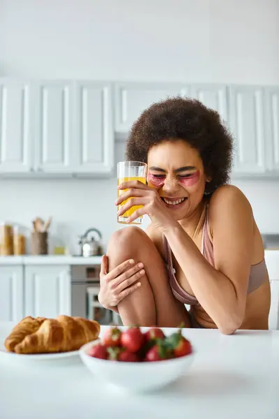 Felice donna africana americana in lingerie bere succo d'arancia vicino alla colazione sul tavolo della cucina — Foto stock