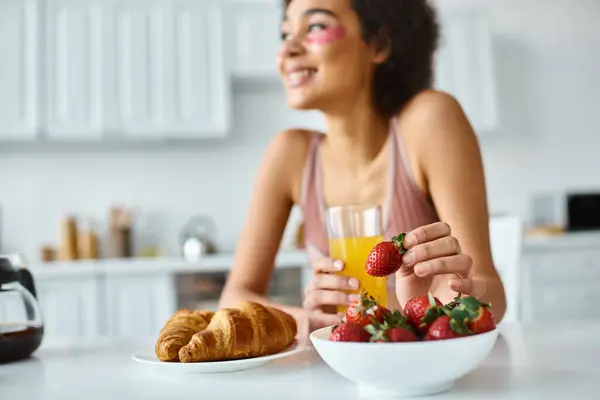 Happy african american woman holding fresh strawberry and orange juice during breakfast at home — Stock Photo