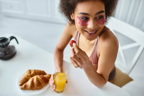 Happy african american woman eating holding strawberry and glass of orange juice in kitchen — Stock Photo