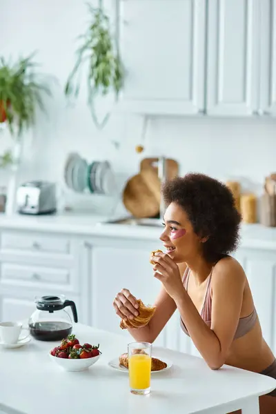 Cheerful african american woman holding freshly baked croissant during breakfast in kitchen — Stock Photo