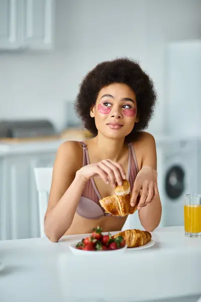 Pensive african american woman holding freshly baked croissant during breakfast in kitchen — Stock Photo
