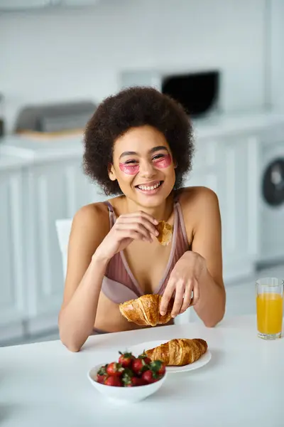 Joyful african american woman holding freshly baked croissant during breakfast in kitchen — Stock Photo