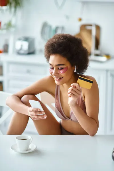 Femme afro-américaine souriante faisant du shopping en ligne avec carte de crédit et smartphone dans la cuisine — Photo de stock
