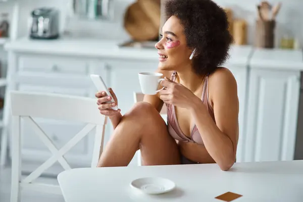 Positive jeune femme afro-américaine tenant une tasse de café et utilisant un smartphone dans la cuisine — Photo de stock