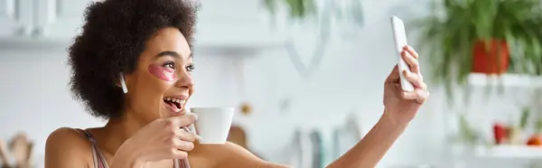 Sonriente mujer afroamericana en lencería con parches para los ojos tomando selfie con taza de café, pancarta - foto de stock