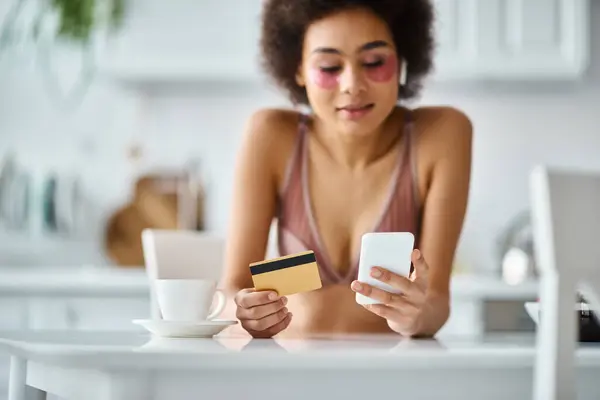 Femme afro-américaine gaie faisant du shopping en ligne avec carte de crédit et smartphone dans la cuisine — Photo de stock