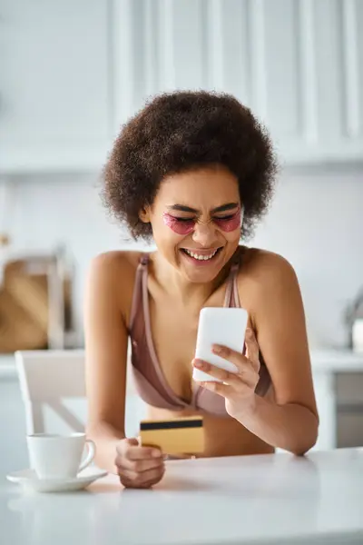 Excité femme afro-américaine shopping en ligne avec carte de crédit et smartphone dans la cuisine — Photo de stock