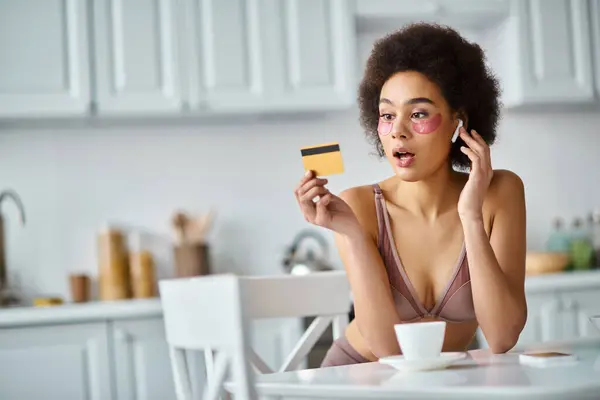 Surpris femme afro-américaine dans des écouteurs avec des patchs sous les yeux en regardant sa carte de crédit — Photo de stock