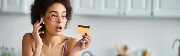Striscione di donna nera sorpresa in auricolare con patch sotto gli occhi guardando la carta di credito — Foto stock