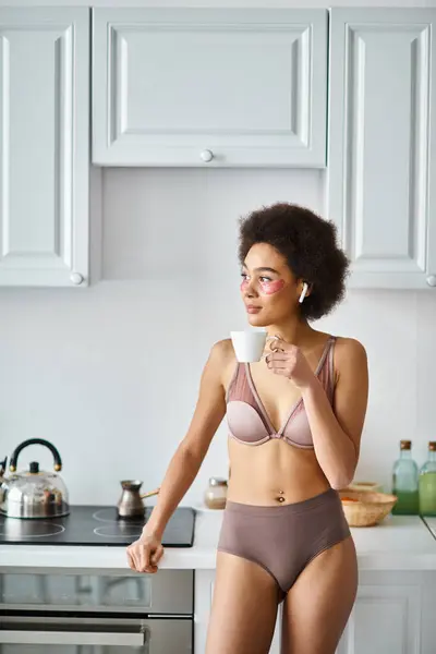 Moment serein avec une femme afro-américaine dans des écouteurs et de la lingerie sirotant le café du matin — Photo de stock