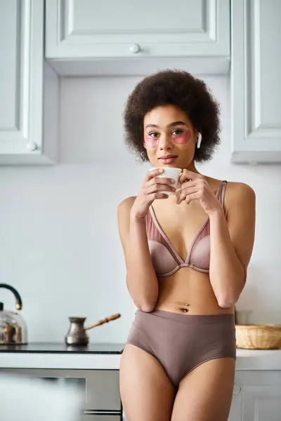 Momento sereno de joven afroamericana en auriculares y lencería bebiendo café de la mañana - foto de stock