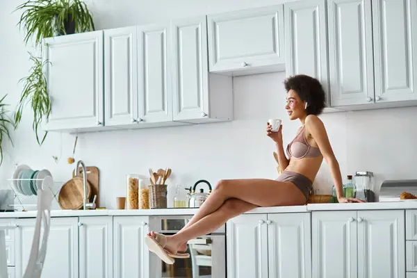 Fröhliche afrikanisch-amerikanische Frau in Dessous, die eine Tasse Kaffee hält und auf einer Arbeitsplatte in der Küche sitzt — Stockfoto