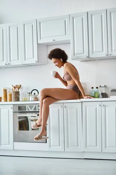 Pleased african american woman in lingerie holding cup of coffee and sitting on kitchen worktop — Stock Photo