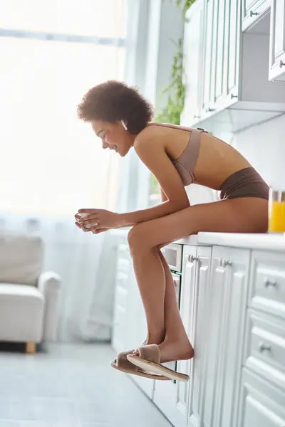 Jolly african american woman in lingerie holding cup of coffee and sitting on kitchen worktop — Stock Photo