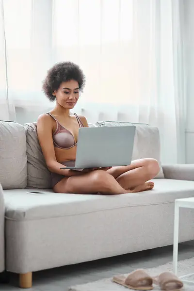 Young african american in lingerie working remotely on laptop while sitting cozy sofa at home — Stock Photo