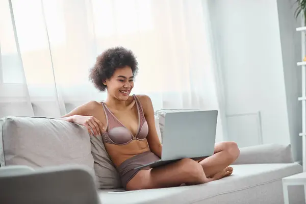 Heureux afro-américaine femme en lingerie assis sur le canapé et regarder le film de comédie sur ordinateur portable — Photo de stock