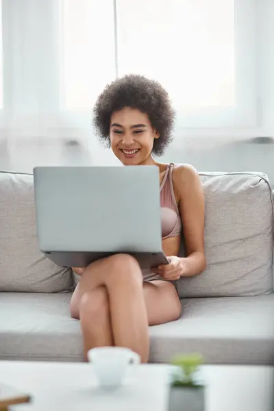 Glücklich afrikanisch-amerikanische Frau in Dessous sitzt auf der Couch und schaut lustige Filme auf dem Laptop zu Hause — Stockfoto