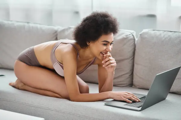 Mujer afroamericana feliz en lencería sentada en el sofá y viendo videos divertidos en el portátil en casa - foto de stock