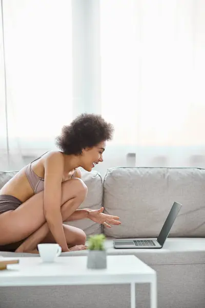Felice donna afroamericana in lingerie seduta sul divano e utilizzando il computer portatile a casa, intrattenimento — Foto stock