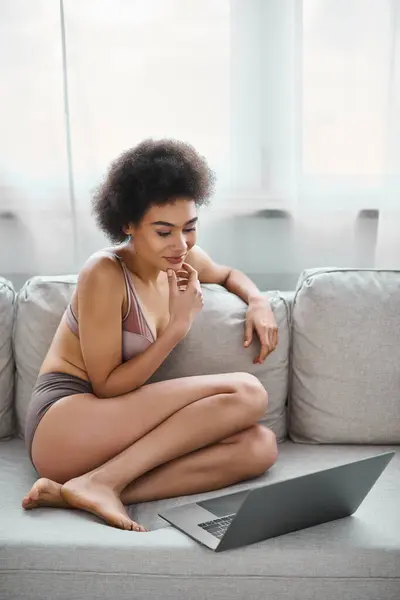 Pensive african american in lingerie working remotely on laptop while sitting on cozy sofa at home — Stock Photo