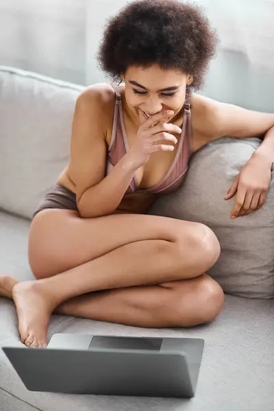 African american woman in lingerie sitting on couch and laughing while watching movie on laptop — Stock Photo