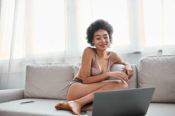 African american woman in lingerie sitting on couch and smiling while watching movie on laptop — Stock Photo
