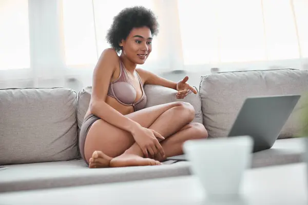 Donna afroamericana in lingerie seduta sul divano e sorridente mentre guarda il computer portatile, fine settimana — Foto stock