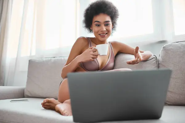 African american woman in lingerie holding cup of coffee and chatting during video call on laptop — Stock Photo