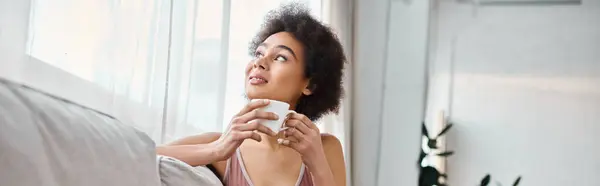 Dreamy african american woman in lingerie holding cup while sitting on sofa at home, banner — Stock Photo