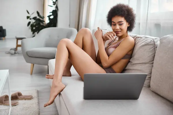 Кучерява афроамериканська жінка в нижній білизні сидить на дивані і дивиться фільм на ноутбуці, вихідні вібрує — стокове фото