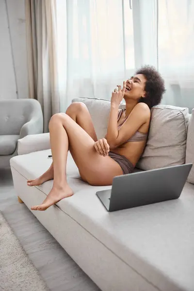 Donna afroamericana in lingerie seduta sul divano e ridendo dal film commedia sul computer portatile, godere — Foto stock