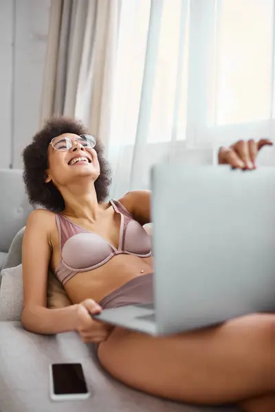 Афроамериканська жінка в білизні в окулярах сидить на дивані і сміється з комедійного фільму — стокове фото