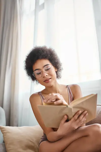 Relaxado e cabelos cacheados afro-americano mulher lendo um livro em lingerie no sofá confortável, sereno — Fotografia de Stock