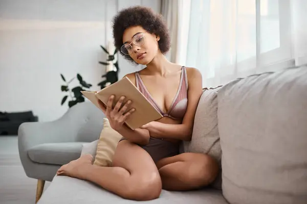 Relaxado e cabelos cacheados afro-americano mulher lendo um livro em lingerie no sofá confortável, sereno — Fotografia de Stock