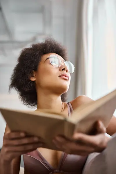 Pensativo e encaracolado Africano americano mulher em lingerie segurando livro e olhando para longe — Fotografia de Stock