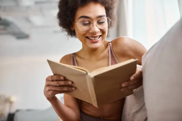 Mulher americana africana alegre e cabelos encaracolados lendo um livro em lingerie em um sofá confortável — Fotografia de Stock