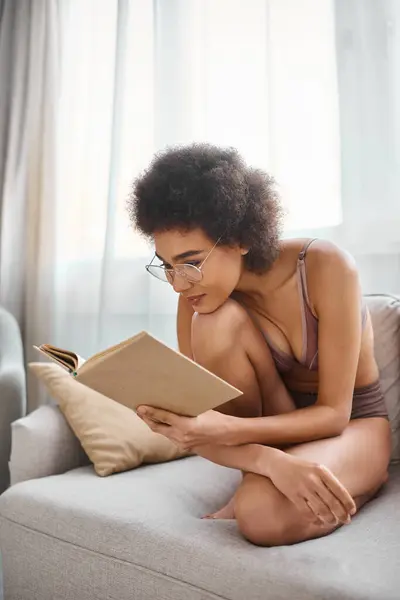Focada e encaracolada afro-americana mulher lendo um livro em lingerie em um sofá confortável — Fotografia de Stock