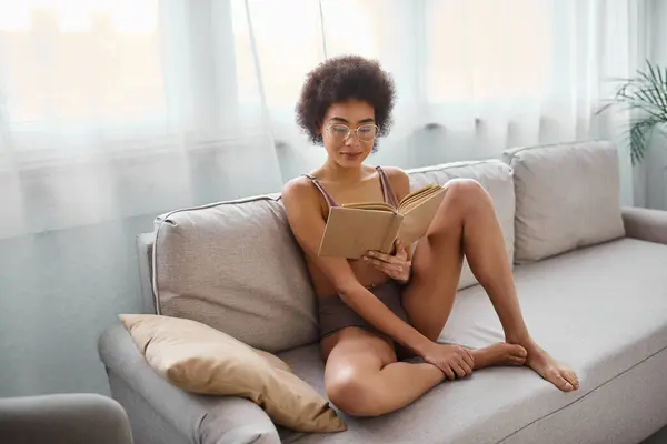 Relaxado e cabelos cacheados afro-americano mulher ler romance em lingerie em um sofá confortável — Fotografia de Stock