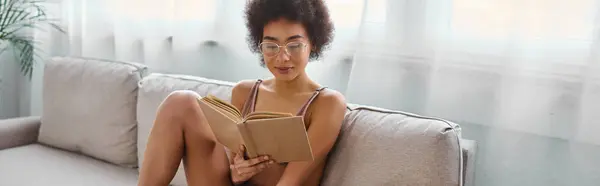 Mujer afroamericana relajada y rizada leyendo un libro en lencería en un cómodo sofá, pancarta - foto de stock