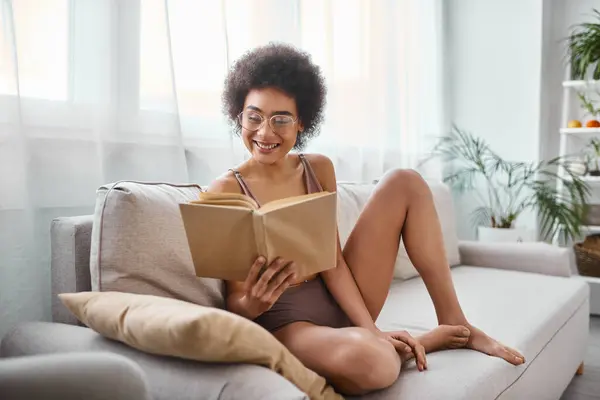 Sorridente e encaracolado afro-americano mulher lendo um livro em lingerie em um sofá confortável — Fotografia de Stock