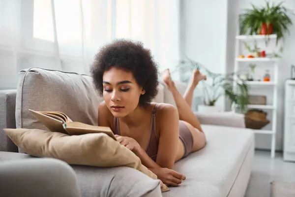 Mujer afroamericana concentrada y rizada leyendo un libro en lencería en un cómodo sofá - foto de stock