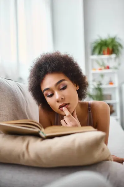 Concentrada e jovem afro-americana lendo um livro em lingerie em um sofá confortável — Fotografia de Stock