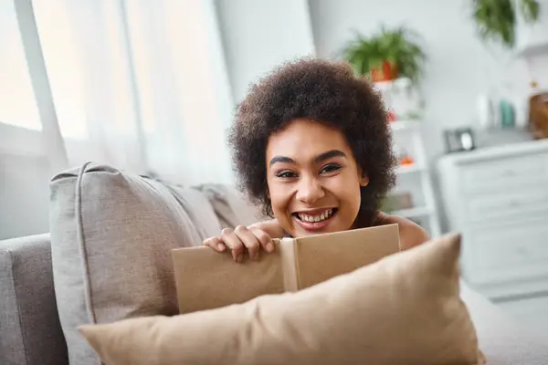Fröhliche und junge afrikanisch-amerikanische Frau, die ein Buch in der Hand hält und zu Hause auf der Couch liegt, genießt — Stockfoto