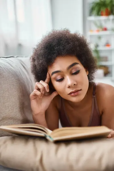 Concentrada e jovem afro-americana lendo um livro em roupa interior em um sofá confortável — Fotografia de Stock