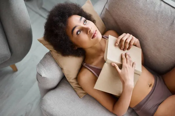 Vista superior da jovem mulher americana africana roupa interior deitado com livro aberto em um sofá confortável — Fotografia de Stock