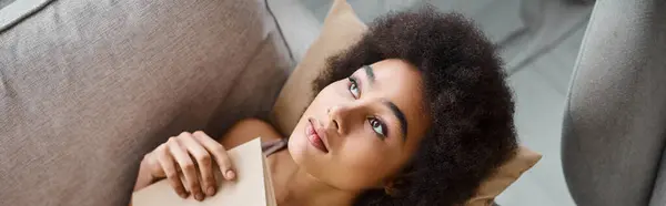 Vista superior da jovem mulher americana africana roupa interior deitado com livro aberto em um sofá confortável, banner — Fotografia de Stock